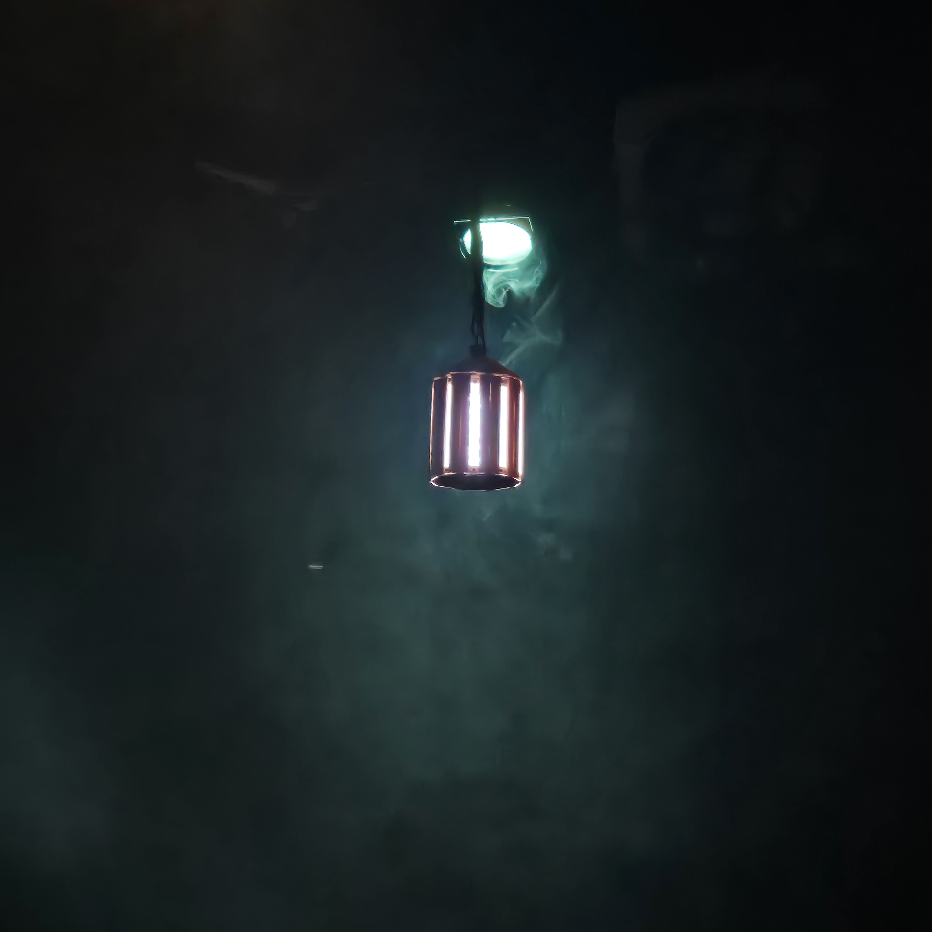 A glowing white lantern hangs in fog turned green by a spotlight above and hehind the lantern.